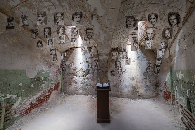 Color photograph of the inside of a windowless, unfurnished cell whose walls are deteriorating to reveal stone and brickwork; fifty sketchbook-size portrait drawings hang from the double barrel-vault ceiling. A pedestal stands in the center of the room with a ledger open toward the viewer.
