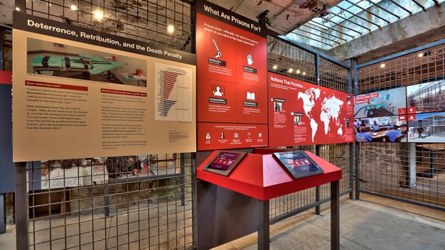 Red graphic panels on wire mesh walls depicting and questioning the main theories of punishment: retribution, deterrence, rehabilitation, and incapacitation.