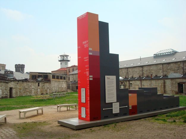 A photograph of the interior of the prison yard containing a massive sculptural installation of a red and gray bar graph displaying data along both axes, set on the ground amidst patches of grass; buildings and the watchtower are seen behind the graph.