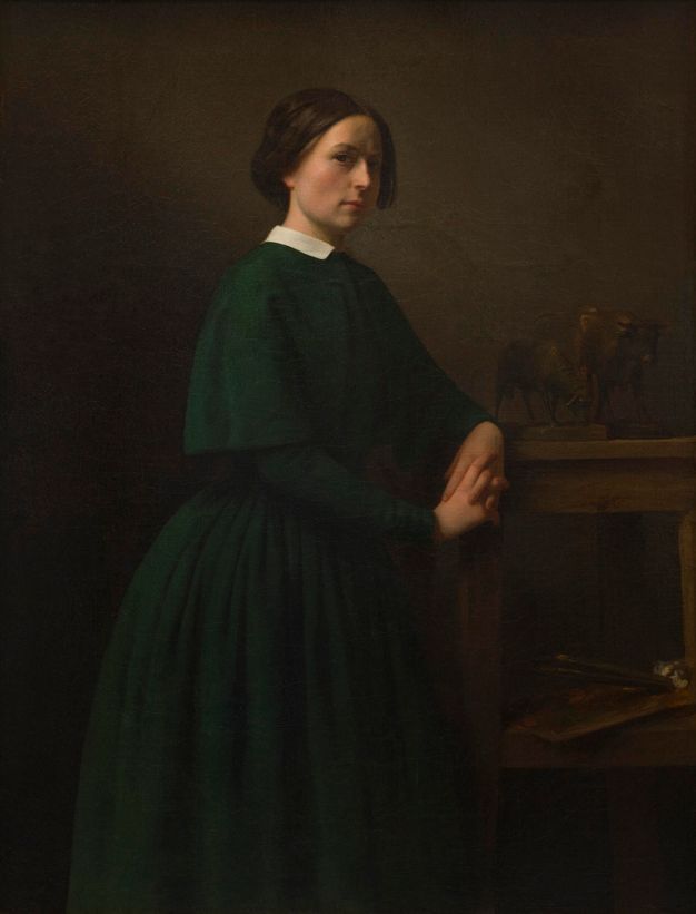 A young woman depicted in three-quarter view in a dark green capelet and dress with a white collar against a dark-brown interior. She leans against a wood table, atop which sit small sculptures of cows; the shelf below holds a palette and brushes.