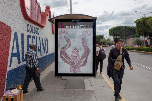 Color photograph of a bus shelter viewed from the middle of a narrow sidewalk along a street, with people walking past. The side pane of the shelter displays Baeza’s name, the exhibition title, and a reproduction of fig. 13. Along the edge of the sidewalk runs a wall painted red, white, and blue.