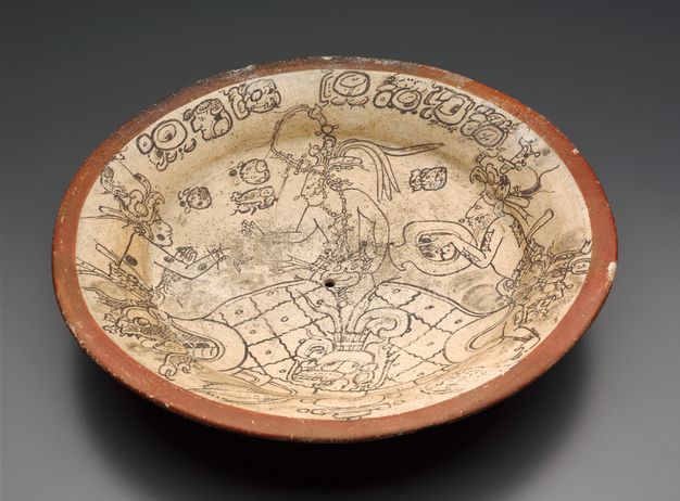 A shallow-rimmed bowl viewed from above. Inside the bowl, which is tan with a red clay–colored rim, three figures carrying vessels are painted in black outline among various vessels and other Maya iconography.