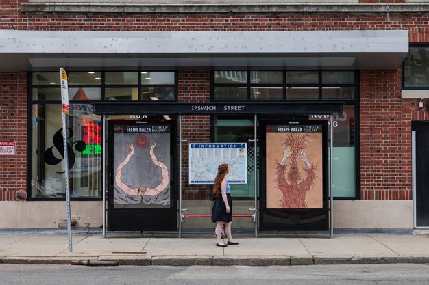 Color photograph of the interior of a bus shelter viewed from across the street, with a woman standing in the center gazing up at one of its two inside panels, which display Baeza’s work, each featuring arm-like forms reaching upward. 
