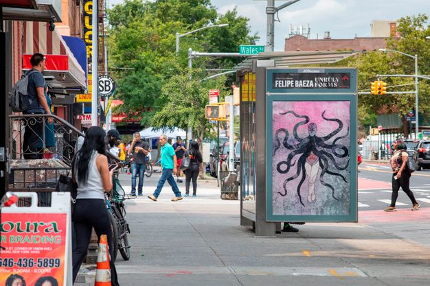 Color photograph of a bus shelter viewed from the side on the large sidewalk of a busy street with many pedestrians walking past along storefronts and building stoops. Baeza’s work is featured on the side panel, featuring a figure with dark tentacle-like limbs against a pink and gray background.