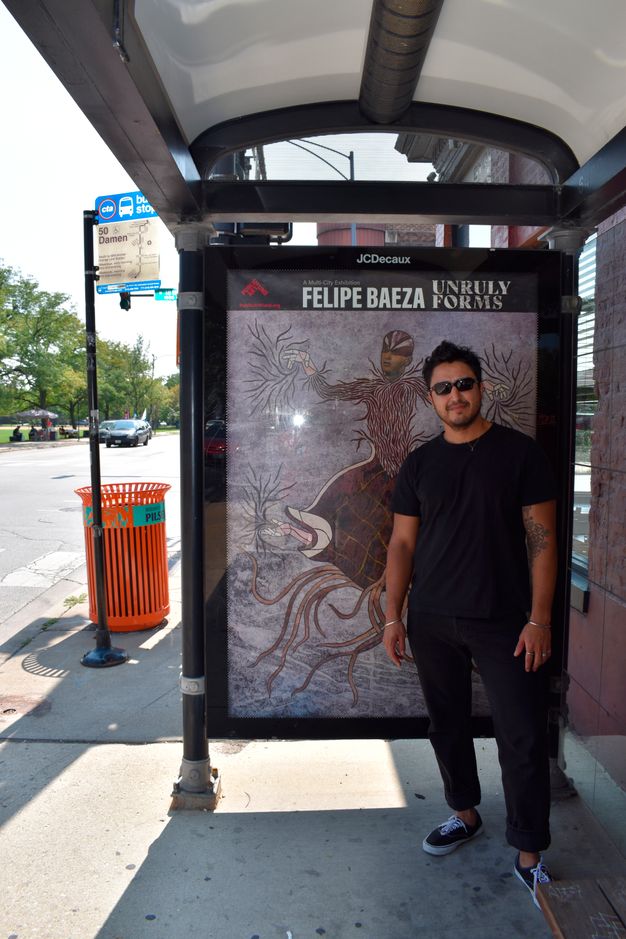 Color photograph of a person slightly smiling toward the viewer under the shade of a bus shelter, wearing sneakers and sunglasses, standing against an interior panel displaying a reproduction of fig. 11. A green park across the street is visible in the background.