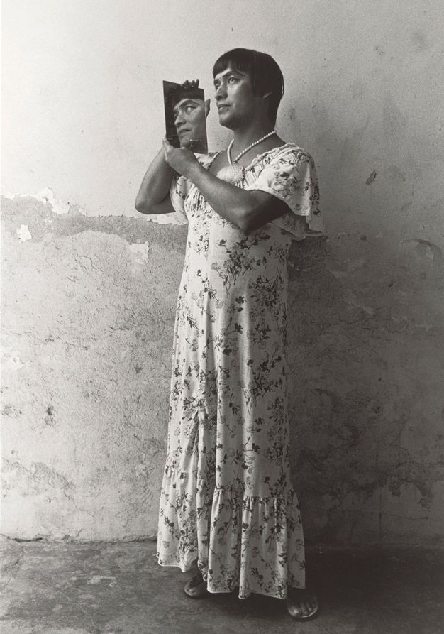 Black-and-white photograph of a person in a standing pose in front of a damaged wall with short, dark hair, wearing a floral dress, sandals, and a necklace, and holding a mirror that reflects their face in profile.