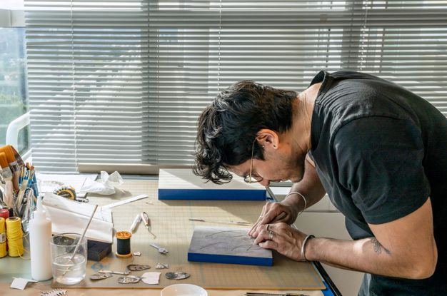 Color photograph of the artist leaning over a worktable wearing a black t-shirt and gold-framed glasses. Art supplies, glues, brushes, and tape measures are scattered around the work surface.