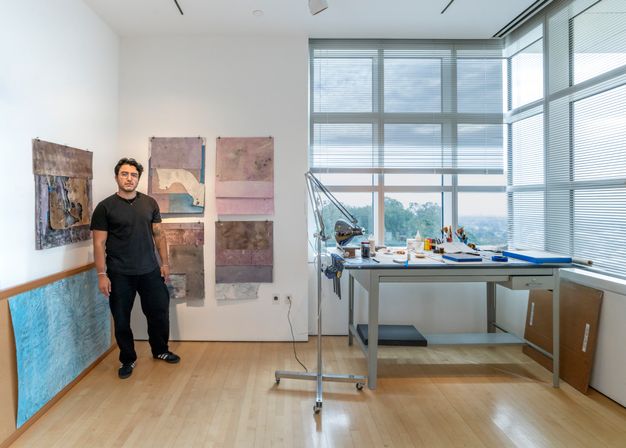 Color photograph of a person standing in the corner of a well-lit art studio in front of works in process tacked to the wall. To the right is a desk with art supplies and corner windows showing a view of Los Angeles.