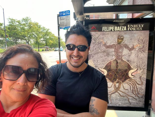 A selfie of two colleagues gently smiling in sunglasses under the shelter of a bus stop in front of its interior side panel, which features a reproduction of fig. 11.