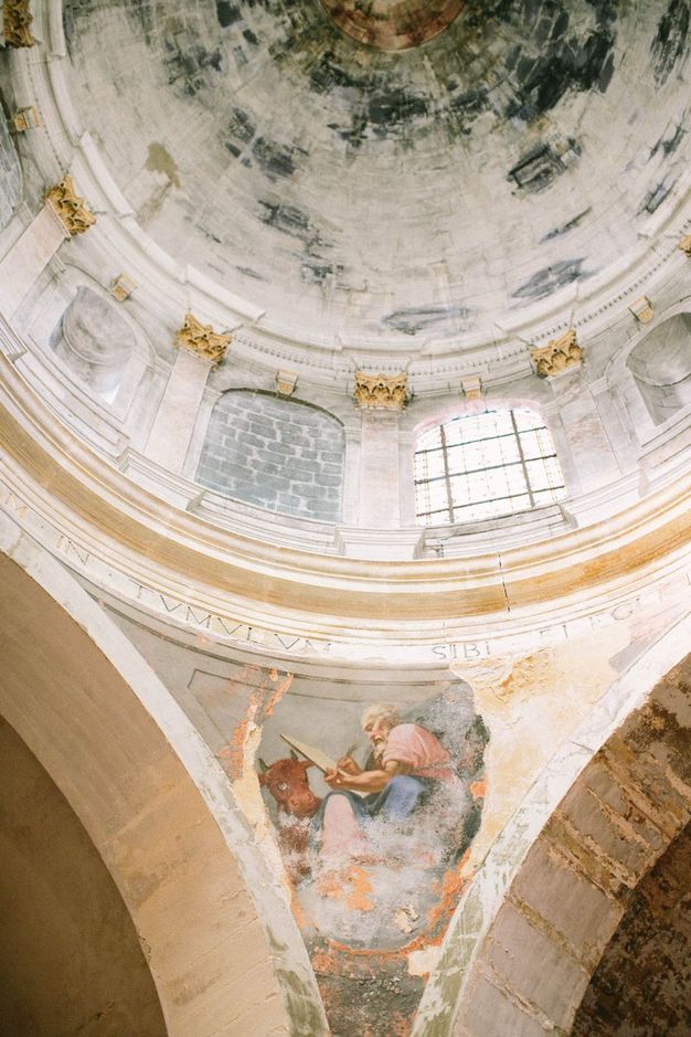 A painted pendentive beneath a dome, as viewed from below. The triangular space depicts a muscular older man in an act of drawing. He wears a loose pink robe and is accompanied by a short-horned reddish-brown cow.