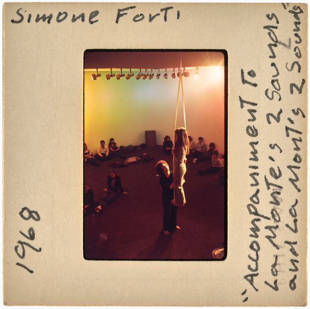 A color slide showing two performers in a carpeted room, one of them suspended midair on a looped rope. Individuals sit and watch along the back wall, onto which colored lights are projected. The frame around the slide contains writing in black ink.