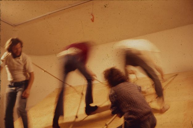 A blurry color photograph of four performers in an interior space climbing an inclined plane of blond wood using knotted ropes.