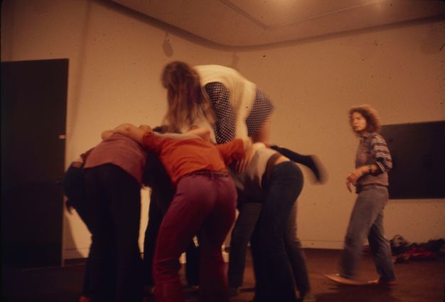 A color photograph of performers huddling in a rounded shape. A long-haired performer in a white vest, a short plaid dress, and knee socks climbs over the backs of the other performers. The choreographer walks around and supervises.