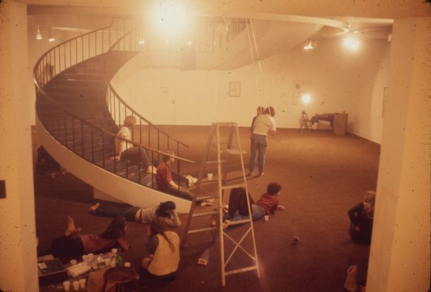 A color photograph of performers sitting on and around a curving staircase in a carpeted museum gallery, watching two people install a rope that hangs down from the ceiling. A ladder stands in the center foreground.