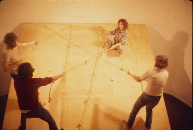 A color photograph of four individuals on an inclined wooden plane. Three performers hold on at the base by the ropes fixed to the top of it. The choreographer holds onto the rope in the upper-right corner, crouching and smiling down toward her colleagues.