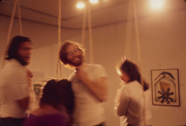 A blurry color photograph of three performers in loops of rope shown from their waists up. The center figure with shaggy hair, a beard, and glasses looks at the camera, tilts his head, and grins.
