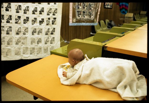 A blanket-wrapped baby lying on its stomach on a table in a laundromat. The baby looks ahead at a large hanging fabric with a photo grid.
