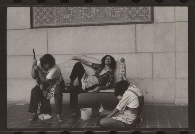 Three women among cleaning supplies. They each sit, slumped over and sprawled out, on a bench or on the ground.