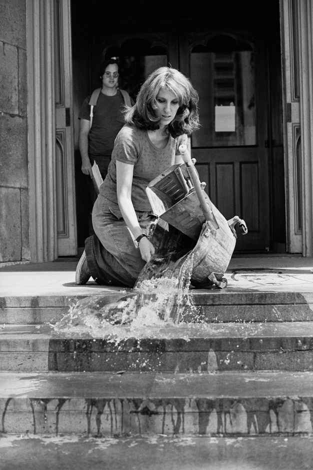 A black-and-white photograph of a woman kneeling and emptying a bucket of water down the steps of a museum, while another looks on from the doorway behind her.