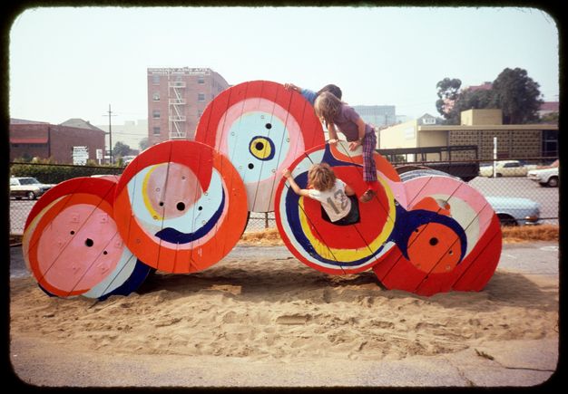 Two children playing on a structure built out of five giant cable spools next to a parking lot. The spools are painted in swirls of bright red, pink, blue, and yellow.