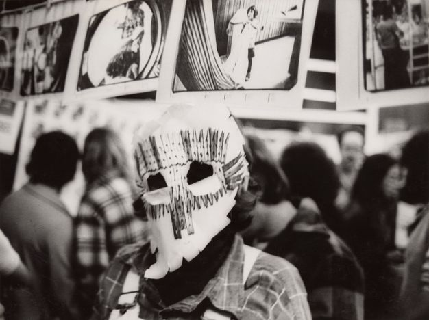 A black-and-white photograph of a masked figure from the shoulders up in a room with artworks strung above on clotheslines. The mask bears a design made out of clothespins.