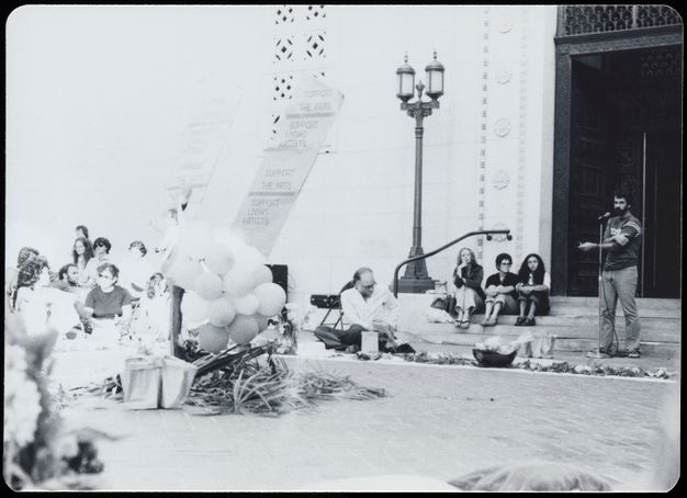 A bearded man speaks at a microphone to an audience gathered outside the entrance of a civic building, next to signs and balloons.