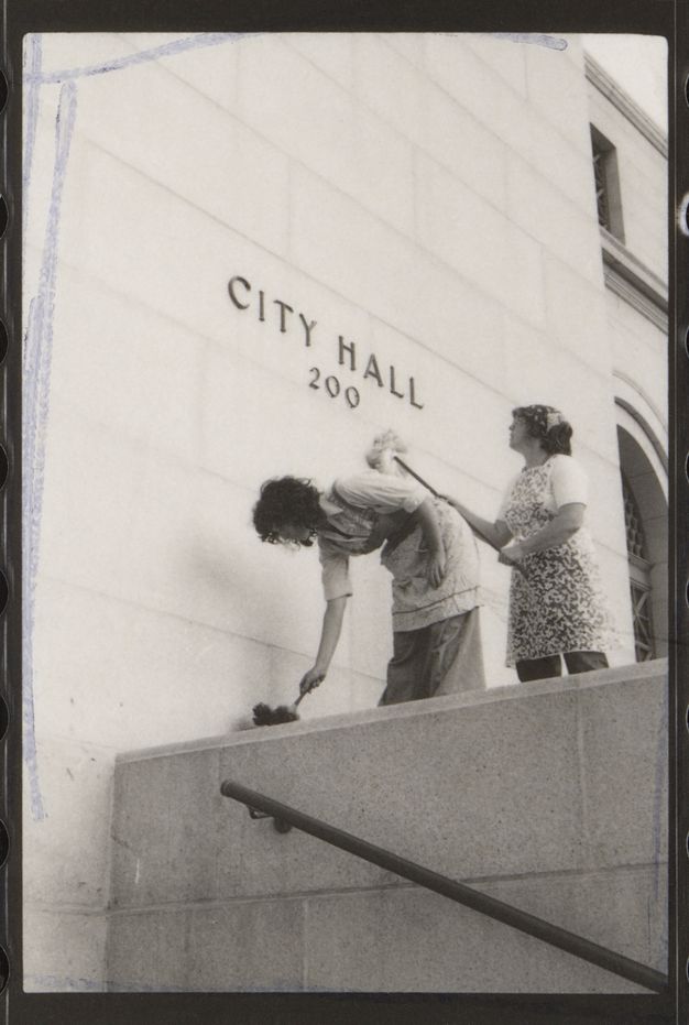 A black-and-white photograph of two women in aprons in the act of washing a facade with mop and duster. The wall bears the words “City Hall, 200.”