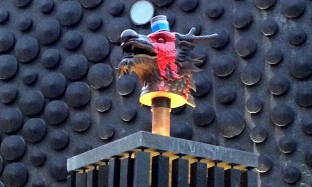 A spilled paint can atop a bronze dragon head. The dragon, with red paint running down the side of its face, over the eye, is mounted on a short pillar installed into a square pedestal.