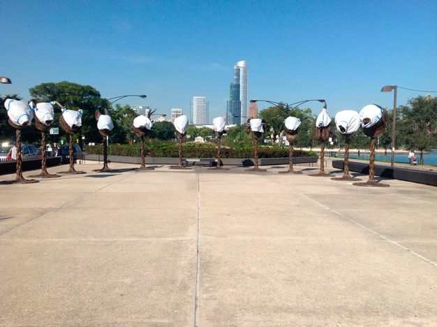 Twelve pillar-mounted animal heads stand in a half-circle at a waterfront park. Each of the heads is wrapped in a white sheet covering the face. Skyscrapers are visible in the skyline behind them.