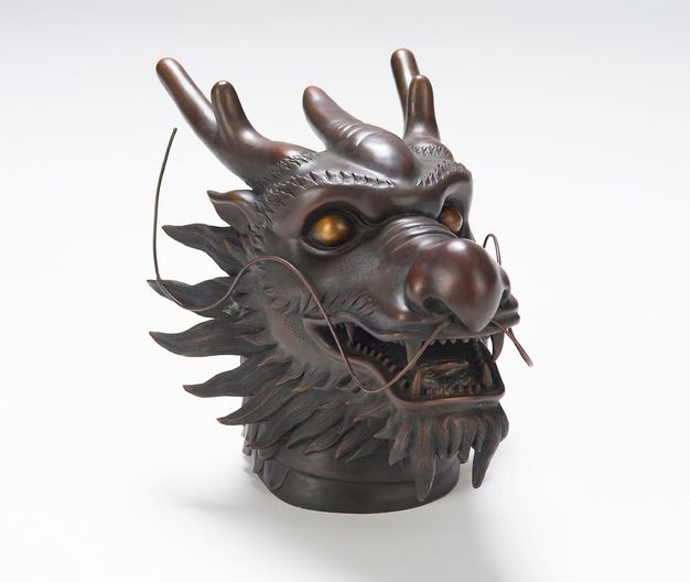 A close-up of a bronze dragon head in three-quarter view against a white background. The dragon’s mouth is slightly open, and a pair of wiry whiskers sweeps back from its bulbous nose.