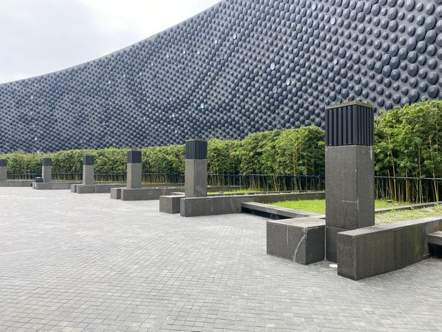 Tall gray rectangular columns line the rounded edge of a paved courtyard. The curved, disc-studded wall of the building behind is lined with hedges.