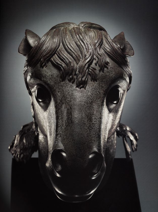 A color photograph of a front-facing bronze horse head seen from slightly above. The sculpture is on a short black pedestal and situated against a dark gray background.