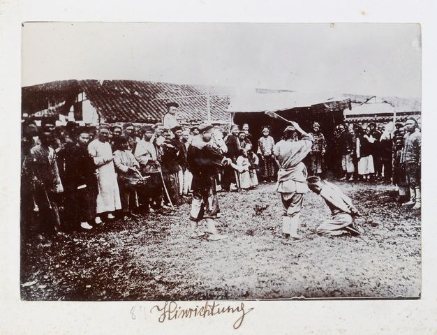 A black-and-white photograph mounted in a book. It shows a man kneeling with bowed head while another man stands over him and raises a sword. A crowd of people stand around them. “Hinrichtung” is written in black cursive below the photo.