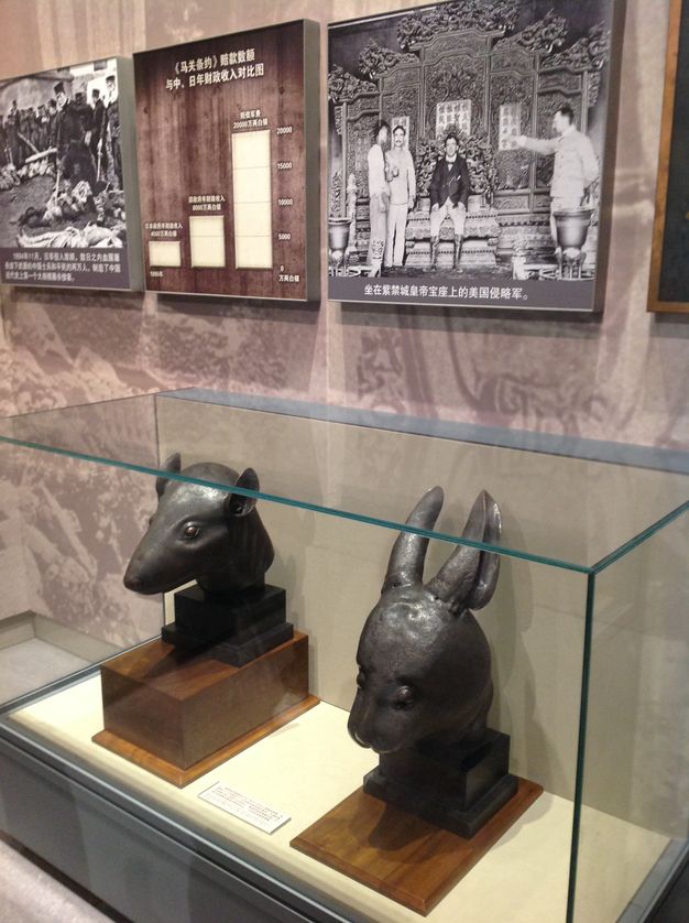 Sculptures of a rat head and a rabbit head inside a display case. The heads are mounted on small wooden pedestals. Black-and-white historic photographs and a bar chart hang on the wall above.