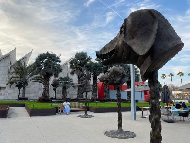 Five towering bronze animal heads in an outdoor area, mounted on thin pillars in a receding arc towards the left. A pig head is the closest in view, followed by that of a dog, rooster, monkey, and ram.
