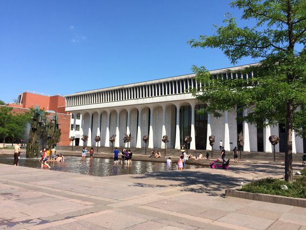 A columned white building with steps leading down to a shallow rectangular fountain. Groups of people stand in the pool or sit on the steps. A row of pillar-mounted animal head sculptures stands between the building and the pool.