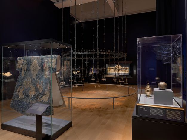 At the center of a dim gallery, an enormous circular chandelier hangs from a metal ring. Suspended from it are metal crosses and trays that once held oil lamps. Around it, display cases hold a silk vestment, precious metal objects, illuminated manuscripts, and a golden altar.
