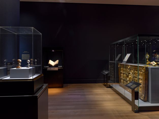 On the left, a display case with a small silver and glass model of an eye accompanied by a gold-tooled leather case faces a display case holding a large, horizontal, rectangular gold panel decorated with relief sculpture showing Christ and scenes from his life, once part of an altar table.