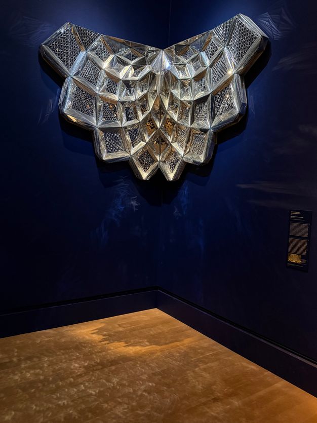 A honeycomb-like, sparkling mirrored sculpture fits into the corner where two dark-blue gallery walls meet. Its surface is covered in mirror mosaic, which fractures the light shining on it and casts refracted light on the walls and floor around it.