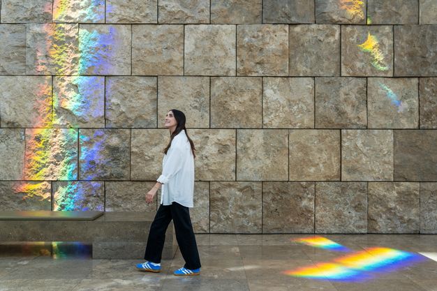A strolling figure admires the bands of spectral light that traverse the textured travertine walls and cast rainbows across the stones and the marble floor.
