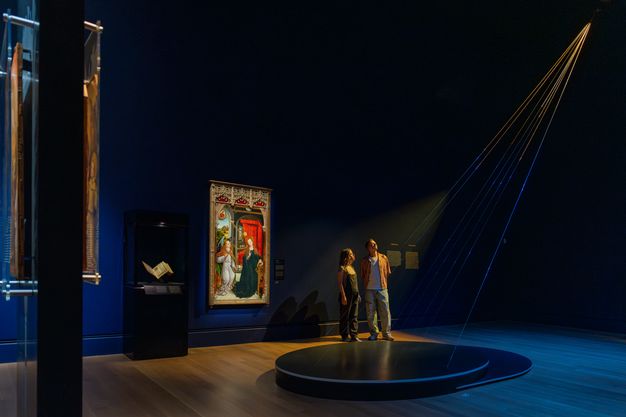 In a gently lit gallery with dark-blue walls, two visitors look up into the light as thin, gilded cables stretch from floor to ceiling, evoking rays of divine light.