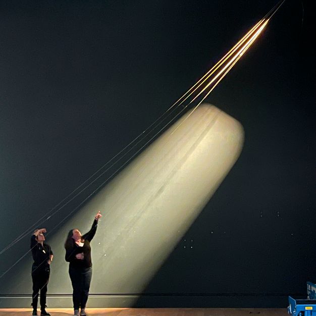 In an empty and darkened gallery, the artist looks up while the lighting designer gestures at thin, gilded cables while spotlights shine from above and along the cables.