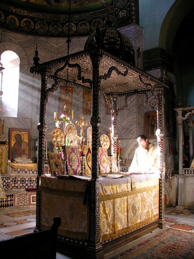 A shaft of early-morning sunlight pierces through the gloom of the bema of a sixth-century church interior. The light gleams on golden liturgical fans and manuscript covers beheld on an ornate wooden altar. A robed individual standing at the altar seems to dissolve in white light.