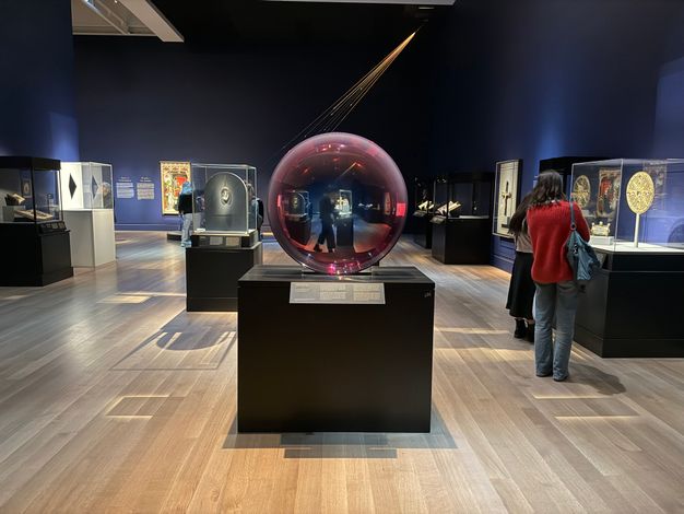 An interior gallery featuring visitors viewing dramatically lit objects in various cases on black mounts. Shadows line the floor. At the center, a reddish, translucent, round sphere whose focal point contains a receding, distorted, and reflected view of a walking figure behind it and the surrounding artwork in cases.