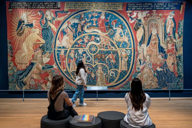 A large, colorful tapestry on the wall of a gallery depicts God emanating light while directing the movement of the cosmos within the circular outline of an astrolabe. Mythological and allegorical figures, including constellations, appear in and around the device.