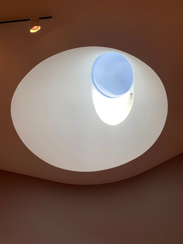 A conical skylight with a round window in a section of the ceiling shows blue sky and an elliptical slice of sunlight.