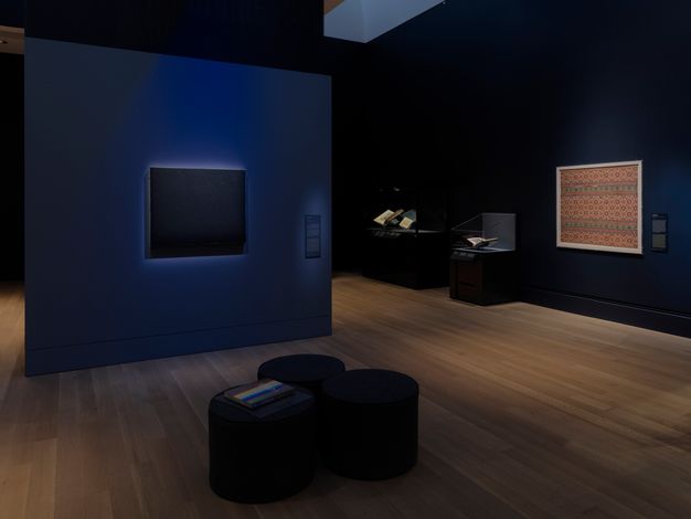 A black canvas with tiny white dots representing stars hangs in low light on a dark-blue wall adjacent to a wall with a similarly sized framed Islamic textile with repeating star pattern.