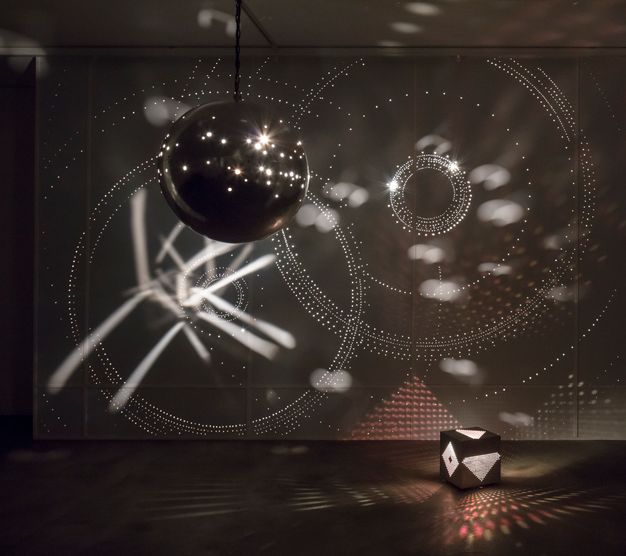 A dark space with a dark, glossy sphere hanging from the ceiling and a cube on the floor, perforated with patterned designs and lit from within. Together they produce geometric light patterns on all surfaces.