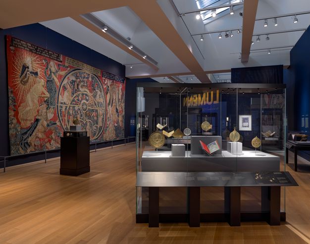 Interior gallery with some natural light containing a massive, brightly colored tapestry on a rear wall depicting an astrolabe and personifications of Philosophy with Geometry, Arithmetic and Astronomy. A transparent display case in the foreground holds astronomical instruments and medieval manuscripts.