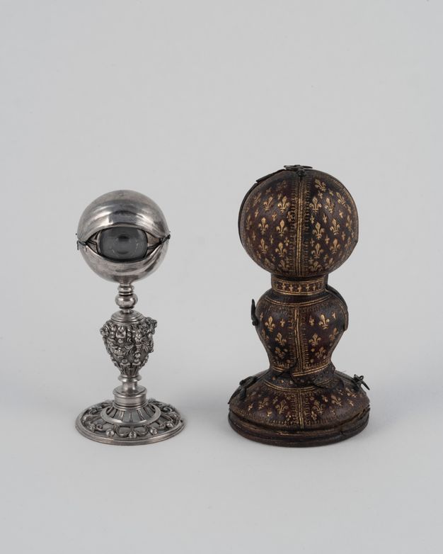 A model of an eyeball on an intricate silver base resembling a candlestick, with a round glass iris hooded in silver, placed next to a gold-tooled leather case with fleur-de-lis designs.